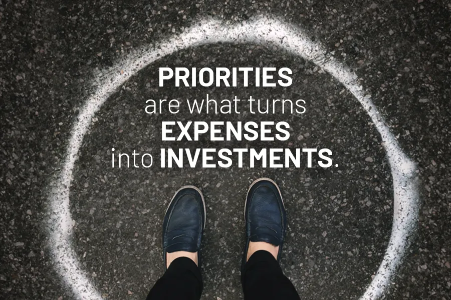 Two feet viewed from above and standing on a pavement circled with the text "PRIORITIES are what turns EXPENSES into INVESTMENTS." overlaying of it.